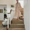 A person wearing a long, light-colored dress walks down a carpeted staircase in a bright, neatly decorated home with white walls, framed artwork, and a stylish seagrass rug.