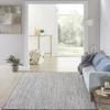 A modern living room with a light blue couch, blue pillows, floor lamp, and gray rug. A plant and pouf sit nearby. Large windows and neutral decor create a bright, airy atmosphere with outdoor isla views of greenery.