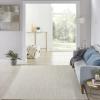 A bright, modern living room with a light rug, pale blue sofa with blue pillows, a floor lamp, plant, and abstract wall art creates a relaxing vibe. Large windows bring in natural light, making it feel like an outdoor isla retreat.