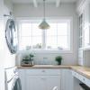 A bright, tidy laundry room with white cabinets, wood countertops, a sink under a window, a stacked washer and dryer, potted plants, a laundry basket in the foreground, and the best rug for laundry room comfort underfoot.